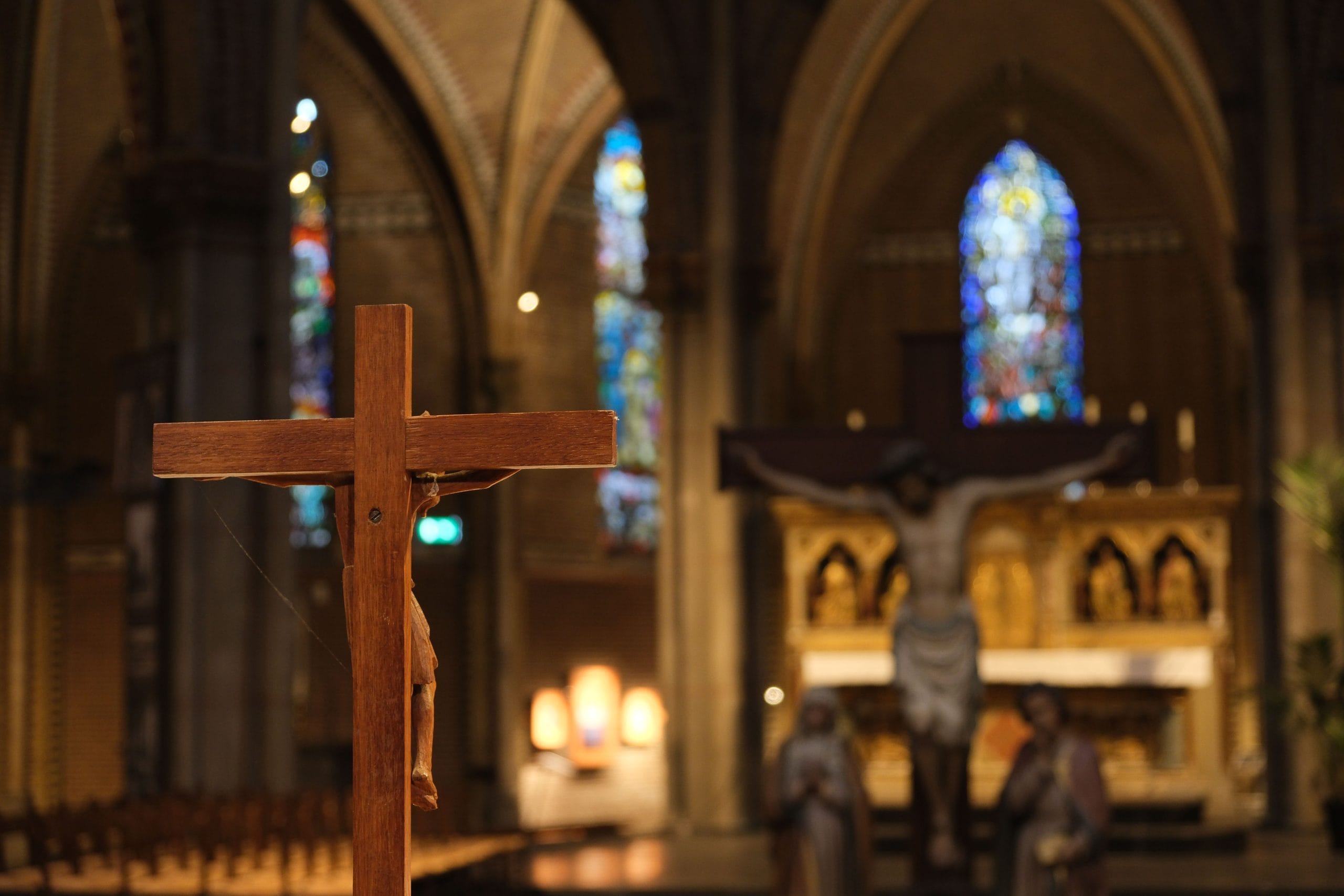 A focused view of a wooden cross in Eindhoven Cathedral with stained glass in the background.
