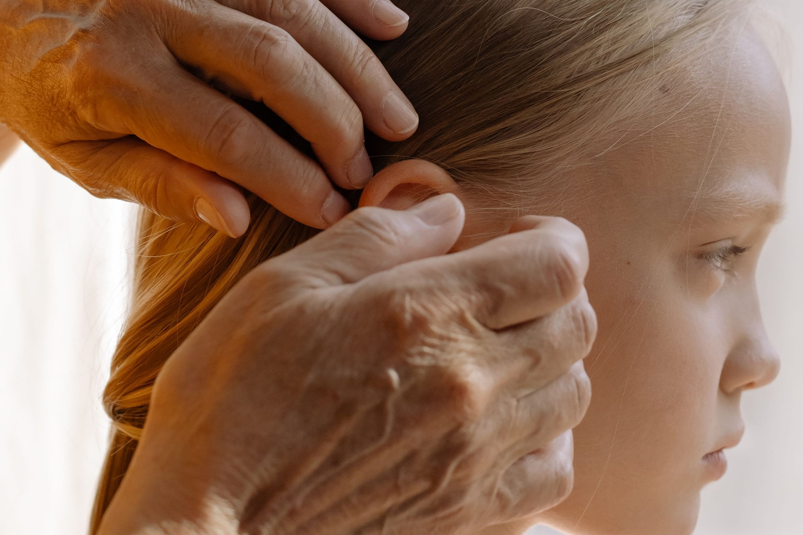 A close-up of an elderly hand arranging a young girl's blonde hair indoors, highlighting multigenerational bond.