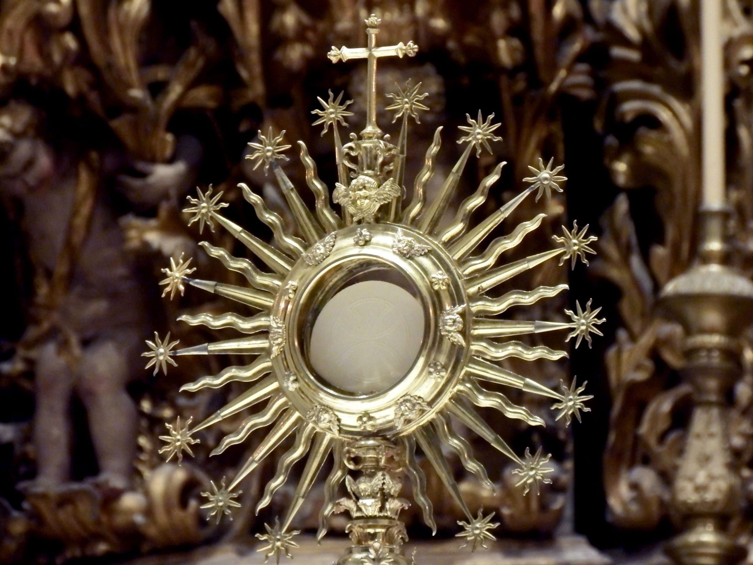 Ornate gilded ostensory in shape of sunburst with Eucharistic host inside against blurred interior of Catholic church