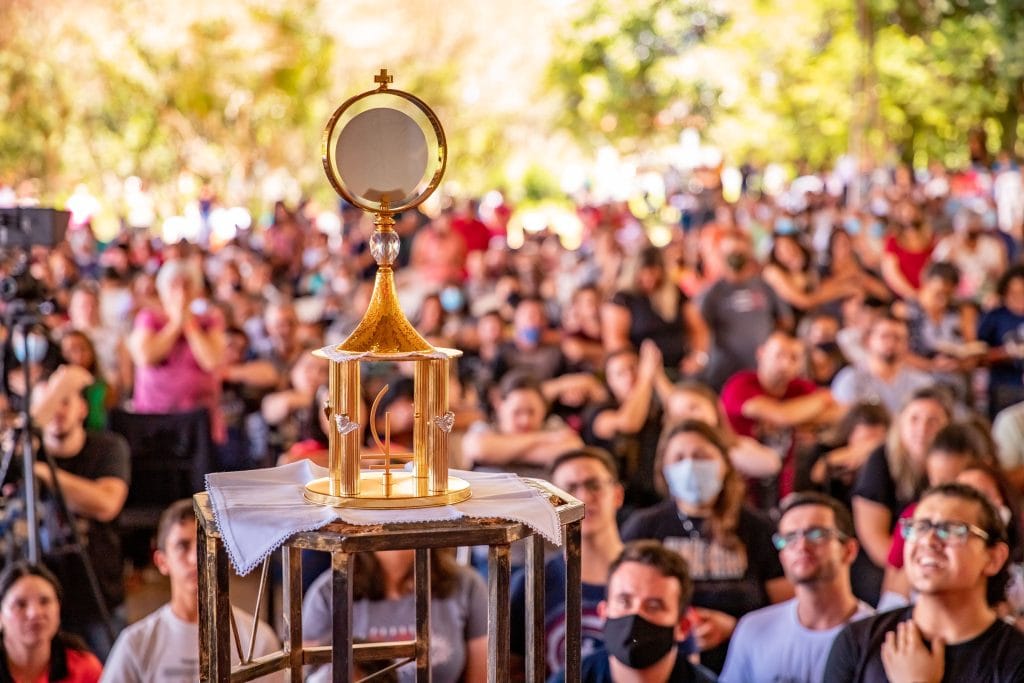 A large crowd attending an outdoor religious ceremony featuring a golden monstrance.